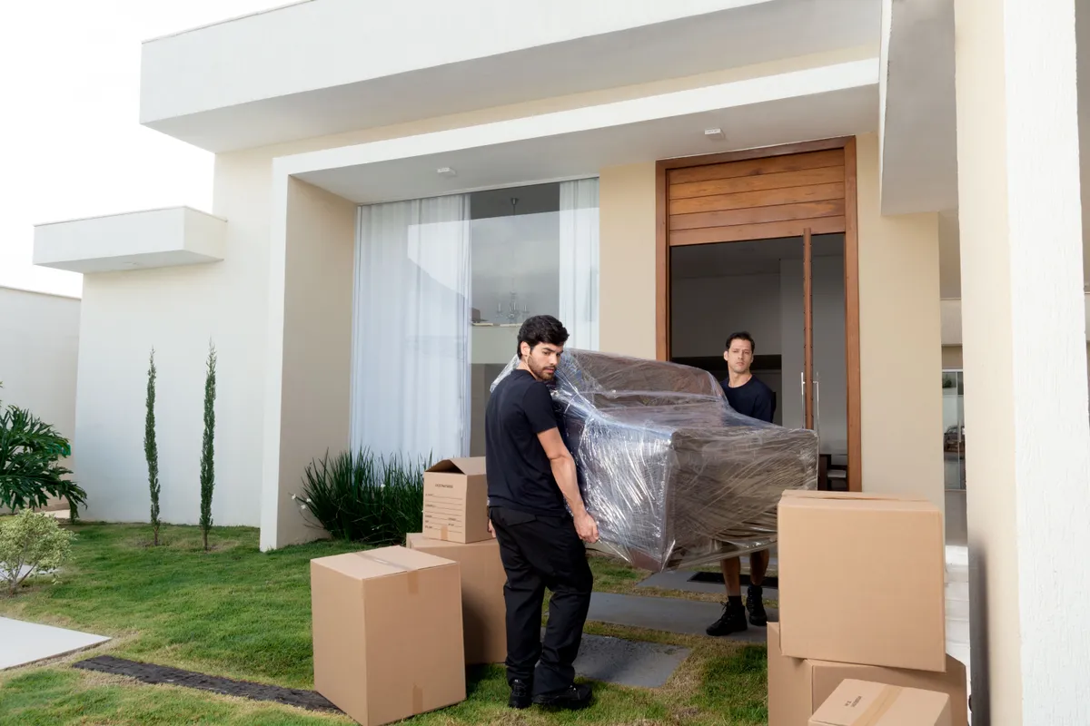 Two service team members carrying a wrapped sofa into a modern home past stacked moving boxes